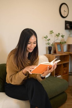 Kahverengi kazak giymiş, oturma odasında kitap okuyan gülümseyen genç bir kadın..