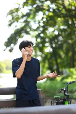 Parkta bisikletinin yanında duran gülümseyen Asyalı adam cep telefonuyla konuşuyor..