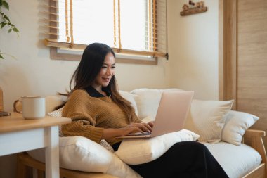 Sıcak kahverengi bir kazak giyen mutlu genç bir kadın kanepede dizüstü bilgisayarla oturuyor..