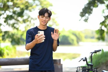 Arka planda göl olan manzaralı bir parkın yanında duran genç adam görüntülü konuşma ya da sanal konuşma yapıyor..