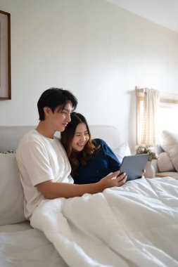 Kaygısız Asyalı çift rahat bir yatak odasında dijital tablet kullanıyor..