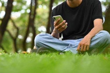 Parkta çimlerin üzerinde otururken akıllı telefon kullanan kırpılmış genç bir adam..