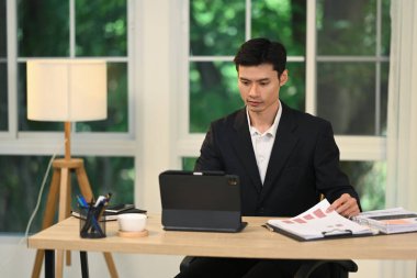 Modern ofise odaklanmış işadamı bir klasördeki belgeleri inceliyor ve dijital tablet kullanıyor.