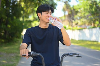 Bisiklete binmeye ve şişeden su içmeye ara veren gülümseyen genç adam..