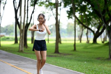 Gülümseyen genç bir kadın parkta koşarken spor bilgilerini kontrol ediyor..