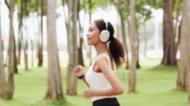 Gülümseyen Afrikalı Amerikalı kadın kulaklık takıyor manzaralı bir parkta koşuyor..