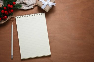 Tatil dekorlu mevsimlik çalışma alanı. Boş defter, kalem, çam dalları ve ahşap masada küçük bir hediye..