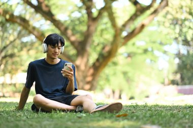 Genç adam müzik dinliyor ve güneşli bir parkta çimlerin üzerinde cep telefonu kullanıyor..