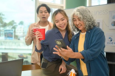 Group of cheerful colleagues enjoying drink and looking at smartphone during an office party. clipart