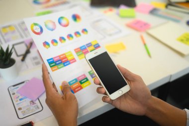 Male UX designer holding smartphone and color palette guide sitting at creative workplace.