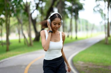 Aktif genç bir kadın yemyeşil bir parkta koşarken kulaklıkla müzik dinlemekten zevk alıyor..