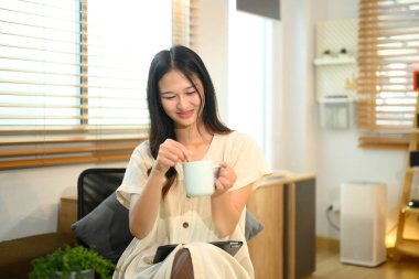 Elinde kahve fincanıyla gülümseyen genç bir kadın. Kanepede dinleniyor. Panjurlardan doğal ışık akıyor..