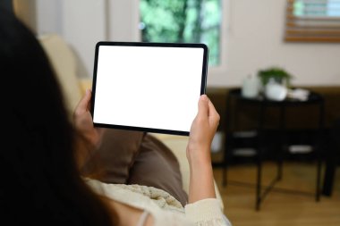 Evdeki kanepede yatarken elinde beyaz ekranla dijital tablet tutan genç bir kadın..