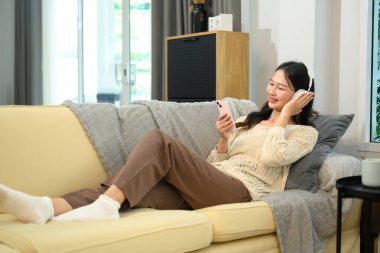 Kablosuz kulaklıklı rahat genç Asyalı bayan kanepede akıllı telefon kullanıyor..