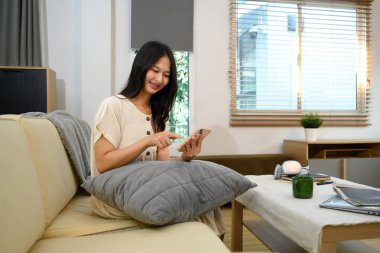 Gülümseyen Asyalı kadın cep telefonuyla mesajlaşıyor şık bir oturma odasında koltukta dinleniyor..