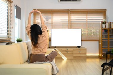 Modern bir oturma odasında kanepeye uzanan genç bir kadın. Tahta konsolda boş TV ekranı var..