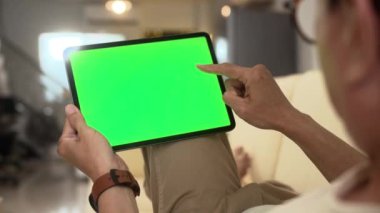 Omzunun üstünden yeşil ekranlı dijital tabletli yaşlı adam evdeki kanepede oturuyor..
