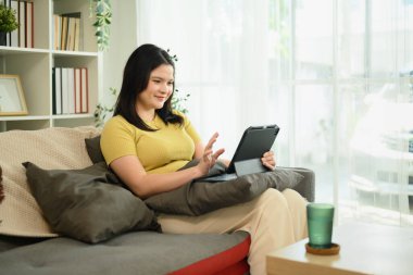 Kanepeye yaslanıp dijital tablette bir şeyler izleyen genç ve mutlu bir kadın..