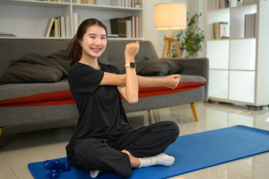 Gülümseyen genç bir kadın yoga minderinde çaprazlama oturuyor ve antrenman yaparken esniyor..