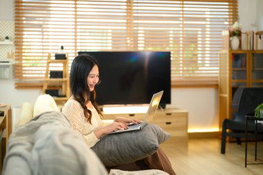 Modern bir oturma odasında dizüstü bilgisayar kullanarak oturan mutlu, genç bir kadın..