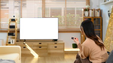 Arka planda elinde uzaktan kumandayla büyük boş bir televizyon izleyen bir kadın var..