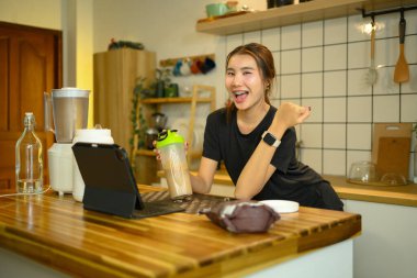 Mutfak tezgahında elinde protein içeceğiyle neşeli genç bir kadın..