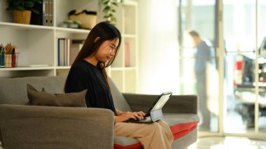 Parlak oturma odasındaki koltukta dijital tablet kullanan gülümseyen genç kadın..