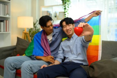 Sevecen eşcinsel çift kanepede oturmuş ellerinde gökkuşağı bayrağı ve kırmızı kalp tutuyorlar..