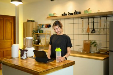 Mutfak tezgahında elinde protein kabıyla dijital tablet kullanan genç bir kadın..