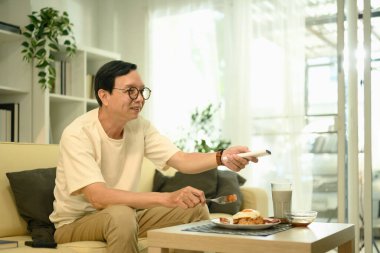 Gülümseyen orta yaşlı bir adam yemek yiyor ve oturma odasında televizyon izliyor..