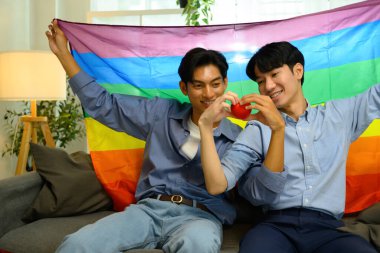 Kırmızı bir kalp taşıyan ve gökkuşağı gurur bayrağı altında oturan neşeli genç erkekler...