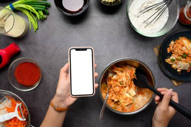 Kimchi 'yi taze malzeme ve baharatlarla çevrili karanlık bir tezgahta hazırlarken el ele tutuşan akıllı telefon..
