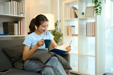 Oturma odasında bir fincan kahve içip kitap okuyan mutlu, genç bir kadın..