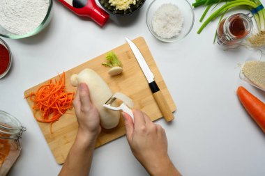 Kimçi malzemeleri ve baharatlarla çevrili ahşap kesme tahtasına daikon turpu soyan eller..