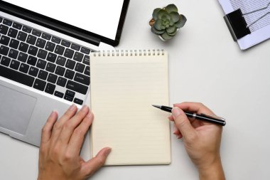 Beyaz masadaki bir dizüstü bilgisayarın üzerinde çalışırken bir adamın spiral bir deftere yazı yazması..