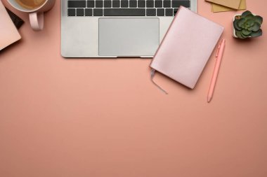 Dizüstü bilgisayarı, defteri, kahve fincanı ve pembe arka planda bitkisi olan üst görünüm kadınsı çalışma alanı.
