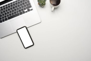 Beyaz ekranlı düz bir akıllı telefon, dizüstü bilgisayar ve beyaz masada bir fincan kahve..