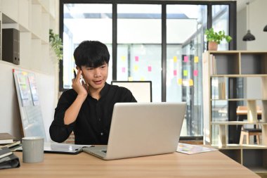 Genç iş adamı iş yerinde dizüstü bilgisayarla telefonda konuşuyor..