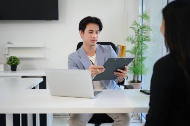 Asyalı erkek işverenin elinde bir pano var. Bir röportaj sırasında adayı dinler ve değerlendirir..