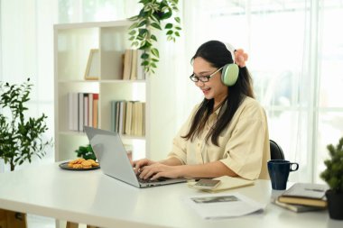 Neşeli, kulaklık takan genç bir kadın. Parlak, rahat bir ofiste dizüstü bilgisayar kullanıyor..