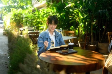 Gülümseyen genç adam, yeşil alanlarla çevrili bir kafede deftere bir şeyler yazıyor...