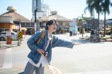 Sarı çantalı ve kameralı gülümseyen bir gezgin şehirde ulaşımı selamlıyor..