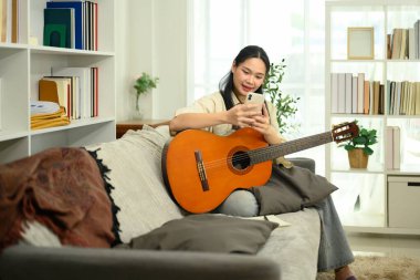 Genç bir kadın rahat bir koltukta gitar çalıyor ve akıllı telefonunu kullanıyor..