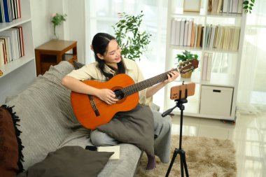 Gülümseyen genç bir kadın gitar çalarken ya da akıllı telefonuyla tripodla kayıt yaparken..