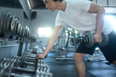 Salonda aynanın önünde duran, kulaklık takan, dambılı kaldıran, odaklanmış bir adamın yan görüntüsü..