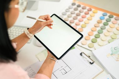 Renk örnekleri, planlar ve tasarım çizimleri ile çevrili dijital tablet üzerinde çalışan tasarımcı.