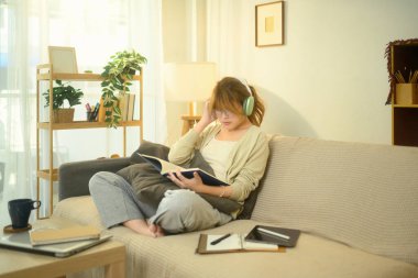 Odaklanmış öğrenci kitap okuyor ve bir oturma odasında kulaklıkla müzik dinliyor..