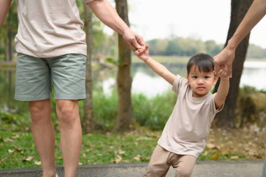 Sevimli ve mutlu küçük çocuk, anne babasının elini tutuyor ve parkta yürüyor..