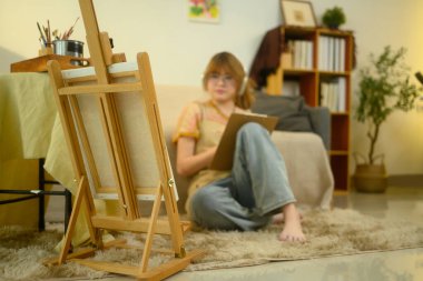 Tabloda resim sehpasında oturan ve çizim defterine odaklanmış kadın ressamlar var..