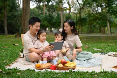 Happy family enjoys picnic in a beautiful park and watching something on digital tablet. clipart